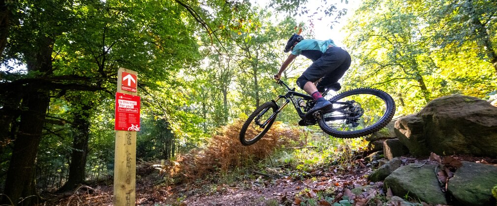 Mountain bike rider doing a jump with a red waymarker sign to his left