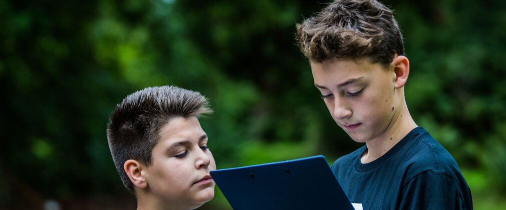 Two boys looking at a clipboard, surrounded by trees.