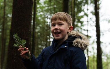 A young boy wearing a winter coat, smiling and holding a pine sprig in the forest.