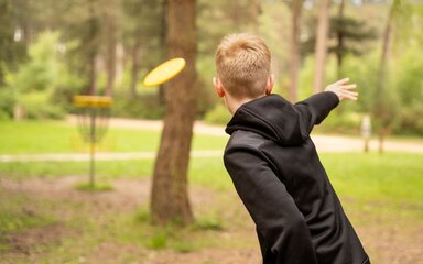Seen from the back, an older boy throwing a yellow flying disc towards a metal basket target.