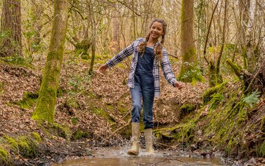 A girl wearing rubber boots splashing in a forest puddle.