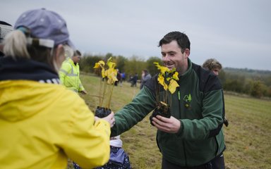 Forestry England worker handing small tree to women in yellow coat 