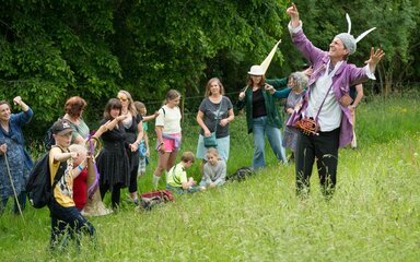 A man in a purple jacket tells an animated story to a group of children and adults in a woodland setting.
