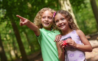 Two smiling young children in the forest. One is pointing at something, the other is holding binoculars.