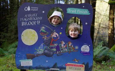 The faces of two children peering through cutouts in a panel in the forest.