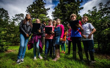 A group of school children, holding clipboards, standing in green space.