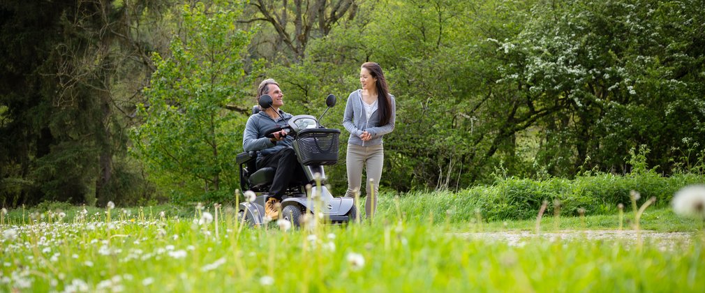A man riding on an all-terrain scooter next to a woman walking through a forest meadow