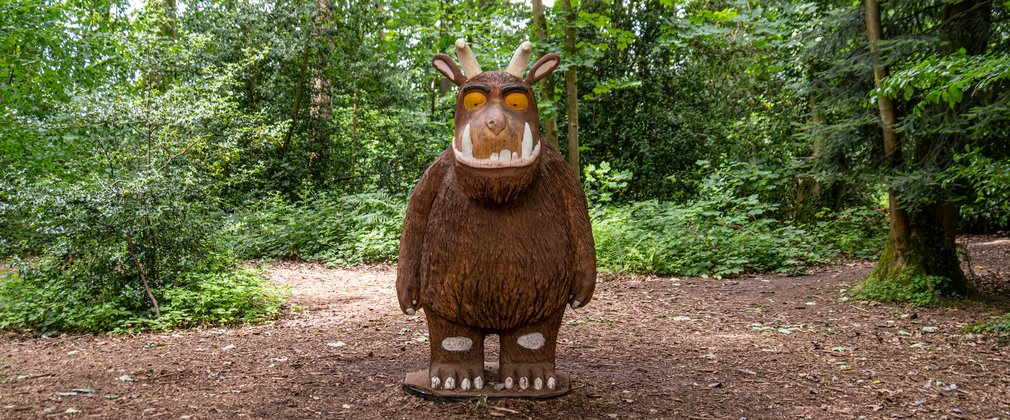 A large wooden sculpture of the children's book character the Gruffalo. Large and Brown with horns, orange eyes and two large fang teeth.