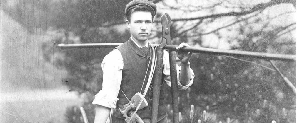 Black and white archive photo of a man wearing a flat cap and carrying gardening tools.
