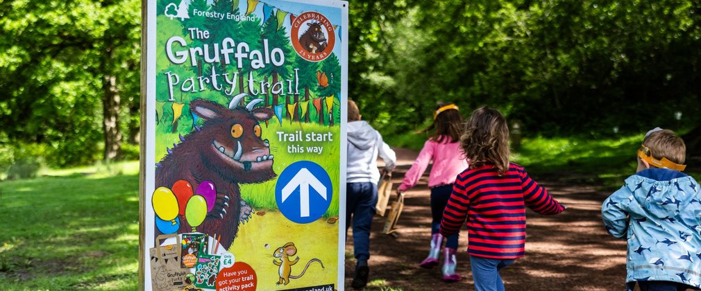 Four children with their backs to the camera walking down a forest path next to a promotional sign for Gruffalo Party Trail