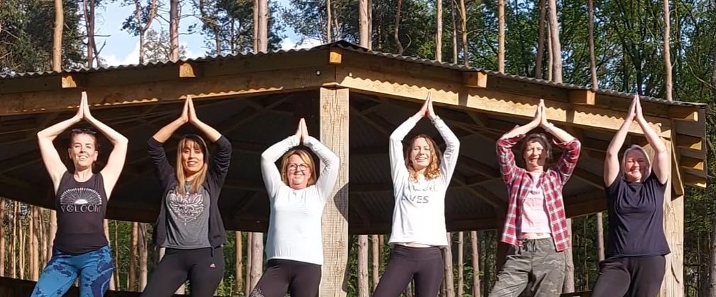 A group of six people standing in 'tree' yoga pose in a forest
