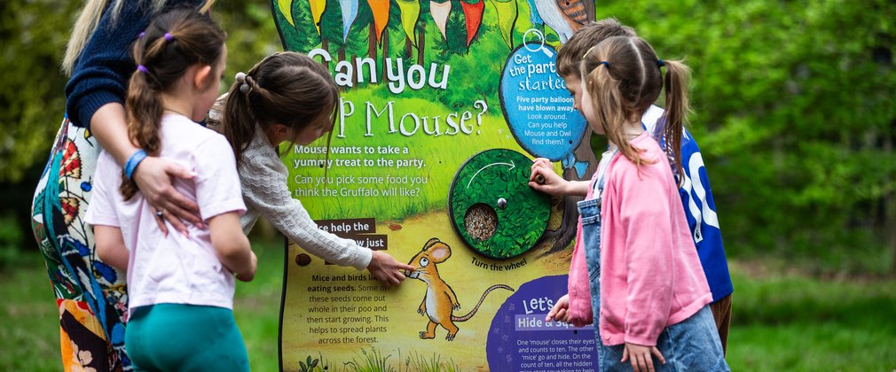4 children crowd round a large Gruffalo panel, they play with the interactive wheel and point to the mouse.