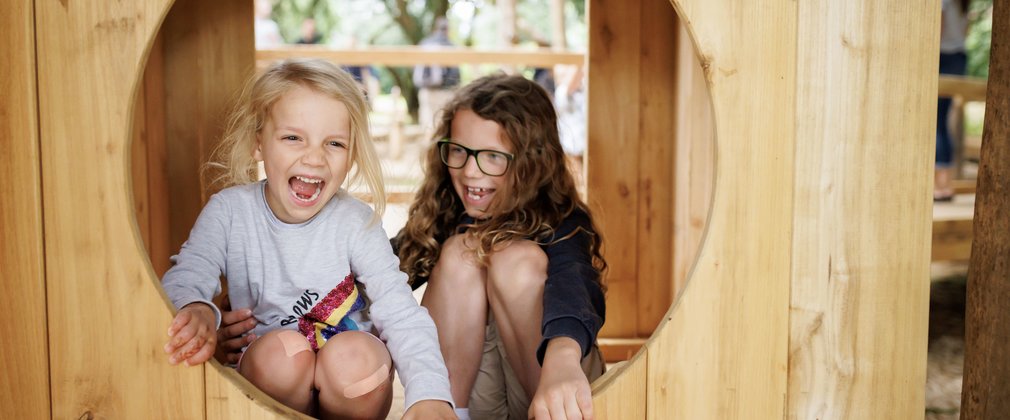 A young girl and boy look cheery through a wooden circular window.