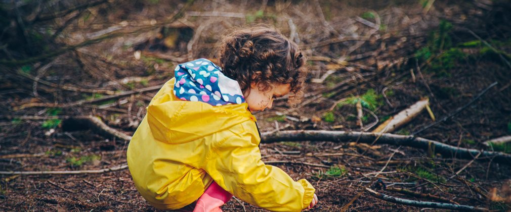 Young girl in yellow coat playing with pine needles on the forest floor