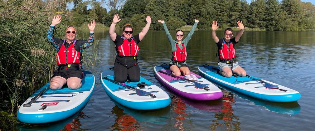 Four people kneeling on paddle boards afloat on water, smiling with their hands in the air.