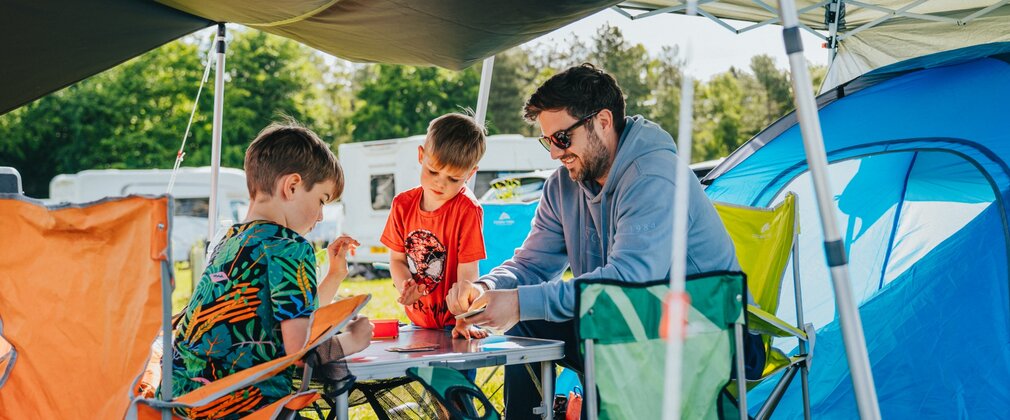 Man and two children sitting around a camping table outside a tent playing cards