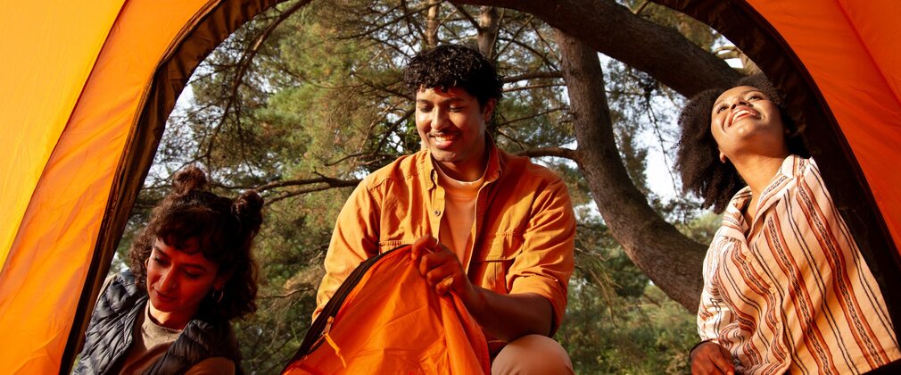 Three people stood outside of an orange tent in the forest