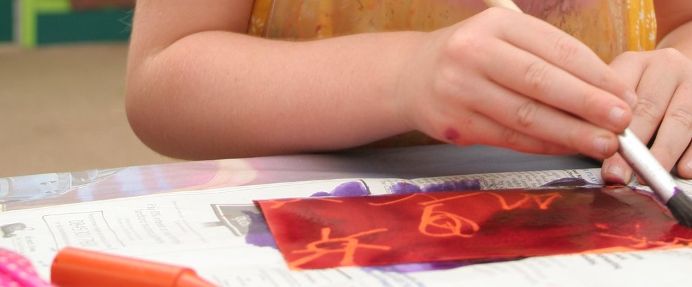 A child holds a paintbrush in her hand. She paints over the piece of paper to reveal a wax drawing of different shapes.