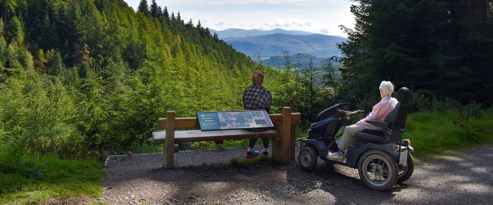 With their backs to the camera, a person sitting on a bench and a person sitting on an all-terrain mobility scooter look out over a high viewpoint