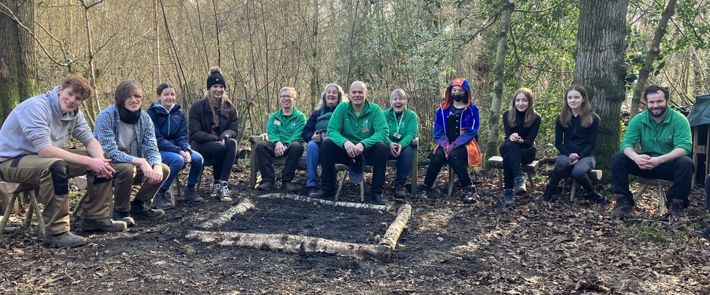 A group of teenagers and adults sit round a square unlit fire pit in the middle of a forest setting, smiling at the camera.