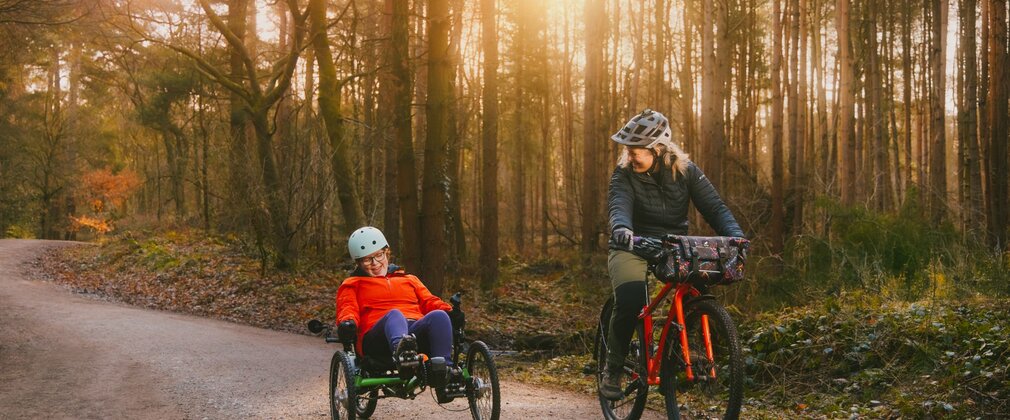 Two women cycling through a forest, one with an adapted bike