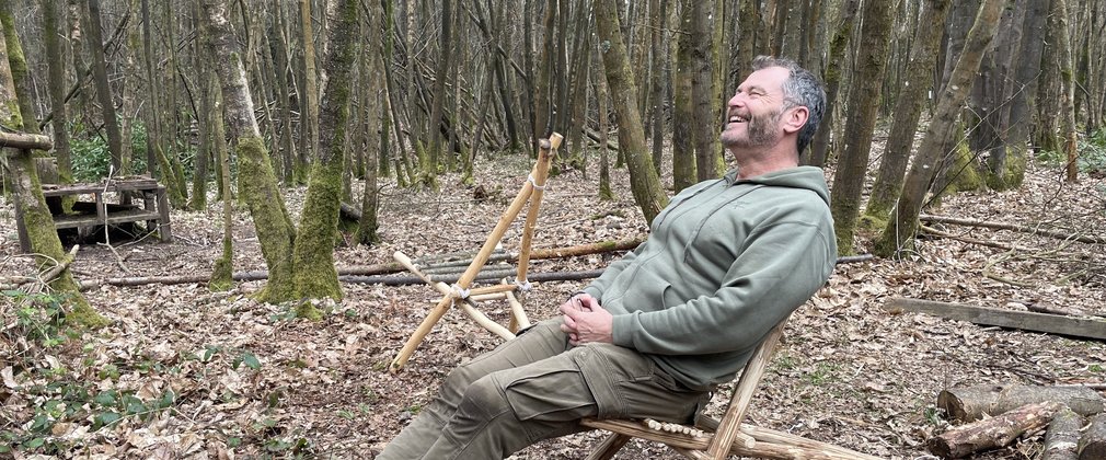 A man sitting back in a homemade wooden chair in a forest setting 