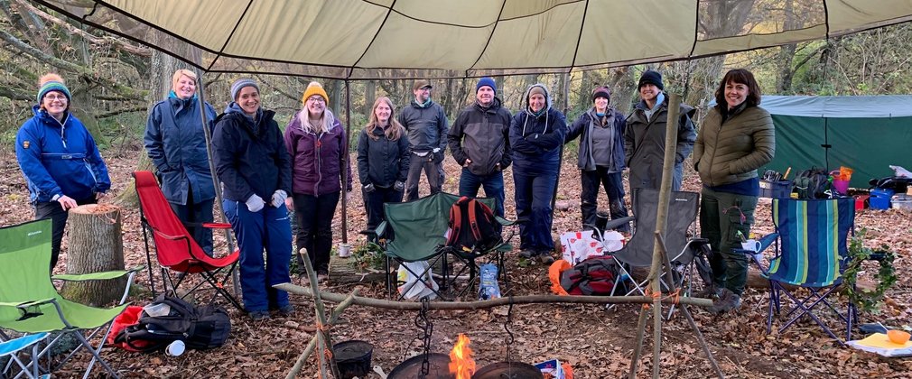 A group of adult forest school students smiling underneath a canopy behind a fire pit 