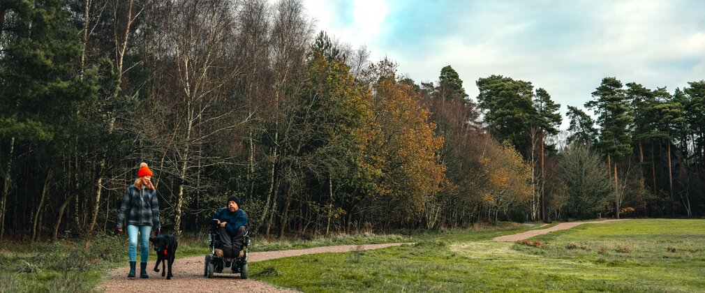 A man using a wheelchair enjoys a leafy trail, with a woman walking a dog