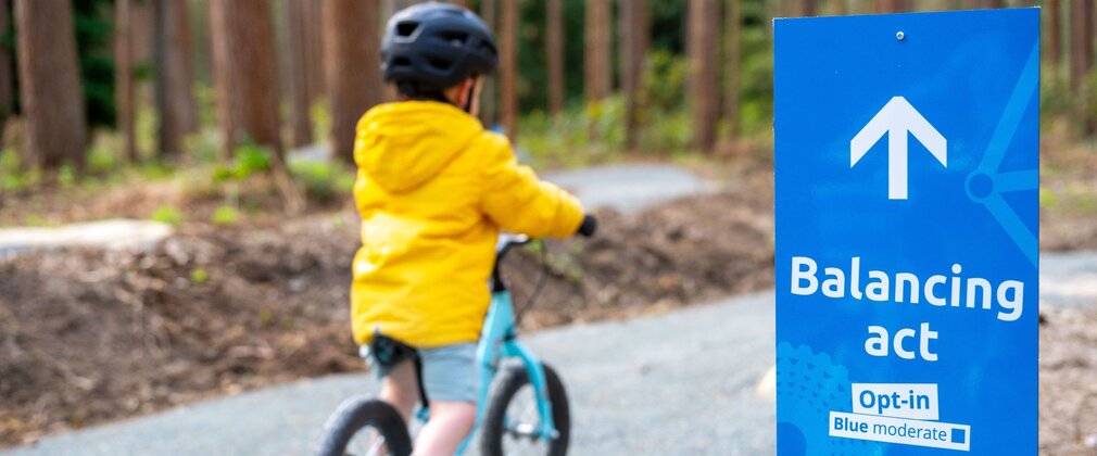Young boy riding a blue balance bike along a forest track.