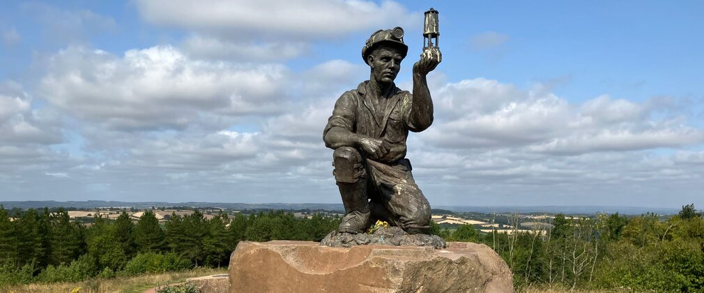 Bronze miner sculpture holding a lamp on a stone plinth at Silverhill Wood, with panoramic countryside views behind.
