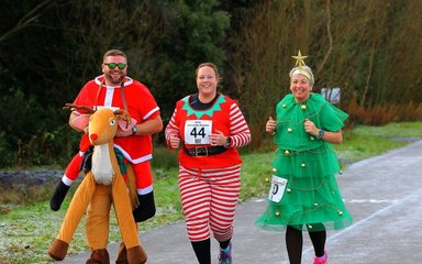 Three people running in a forest dressed as a reindeer, elf and Christmas tree