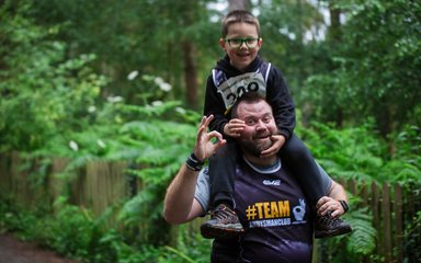 A smiling man in a running top gestures at the camera, with a young boy sat on his shoulders.