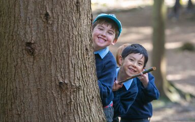 Two young boys in school uniform smile as their peer around a large tree trunk.