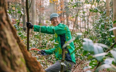 A man wearing a green Forestry England jacket in the woods, smiling at the camera and holding a handsaw and small branch