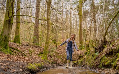 A girl wearing wellington boots splashing in a muddy puddle in the forest, with bare trees and leaves on the ground.