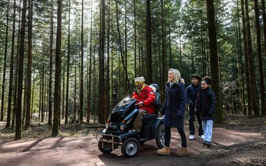 two adults and two children on a forest path, with one adult using an all-terrain mobility scooter.