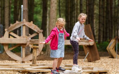 Two young girls balancing on a log in a forest play area