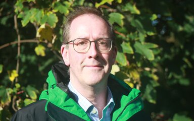 Headshot of a white man wearing glasses standing in front of green foliage.