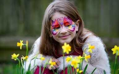 A girl with her face painted with a butterfly looks contently at some daffodils