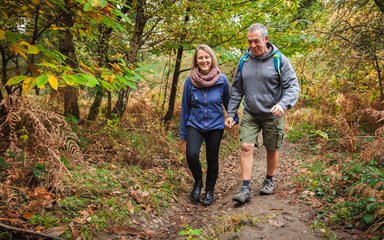 A man and woman smiling and holding hands as they walk on a path surrounded by autumn trees with leaves on the ground.