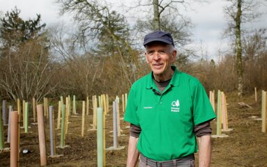 A male volunteer in a green jacket stands smiling in a woodland surrounded by colourful tree tubes