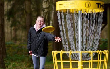 A young person throwing a yellow flying disc towards a disc golf net.