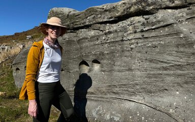 Woman in a hat and mustard cardigan by a large rock.