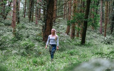 A white woman with long blond hair walking along a forest path surrounded by greenery.