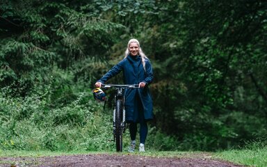 A white woman with long blond ponytail wheels a mountain bike along a forest path.