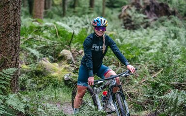 A woman wearing cycling clothes, helmet and sunglasses poses atop a bike in the forest.