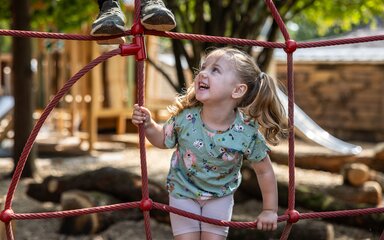 A little girl is climbing a red robe web and smiles as she looks up.