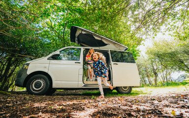 Two girls running out of a white campervan surrounded by trees