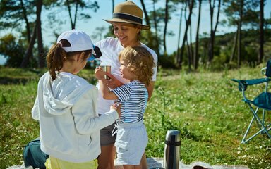 A smiling woman wearing a summer hat pours a drink from a flask, on a picnic blanket with two children.
