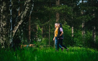 Two women walking side by side in the forest, with a small dog.
